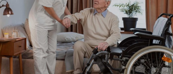 Une soignante aide une personne âgée à se lever, avec un fauteuil roulant à proximité.